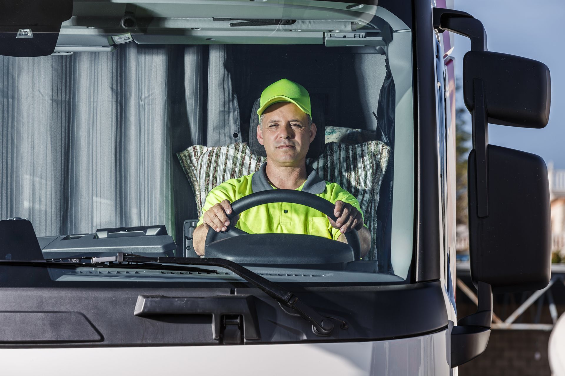 Truck driver wearing a neon vest and hat, sitting in the cab, smiling, holding the steering wheel.