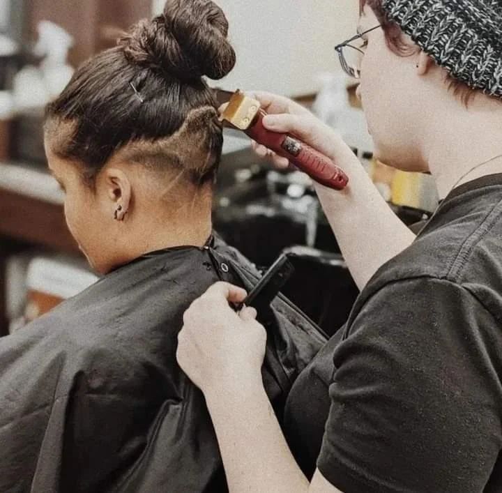 Person getting a haircut, a stylist using clippers to create a design on the nape of their neck.