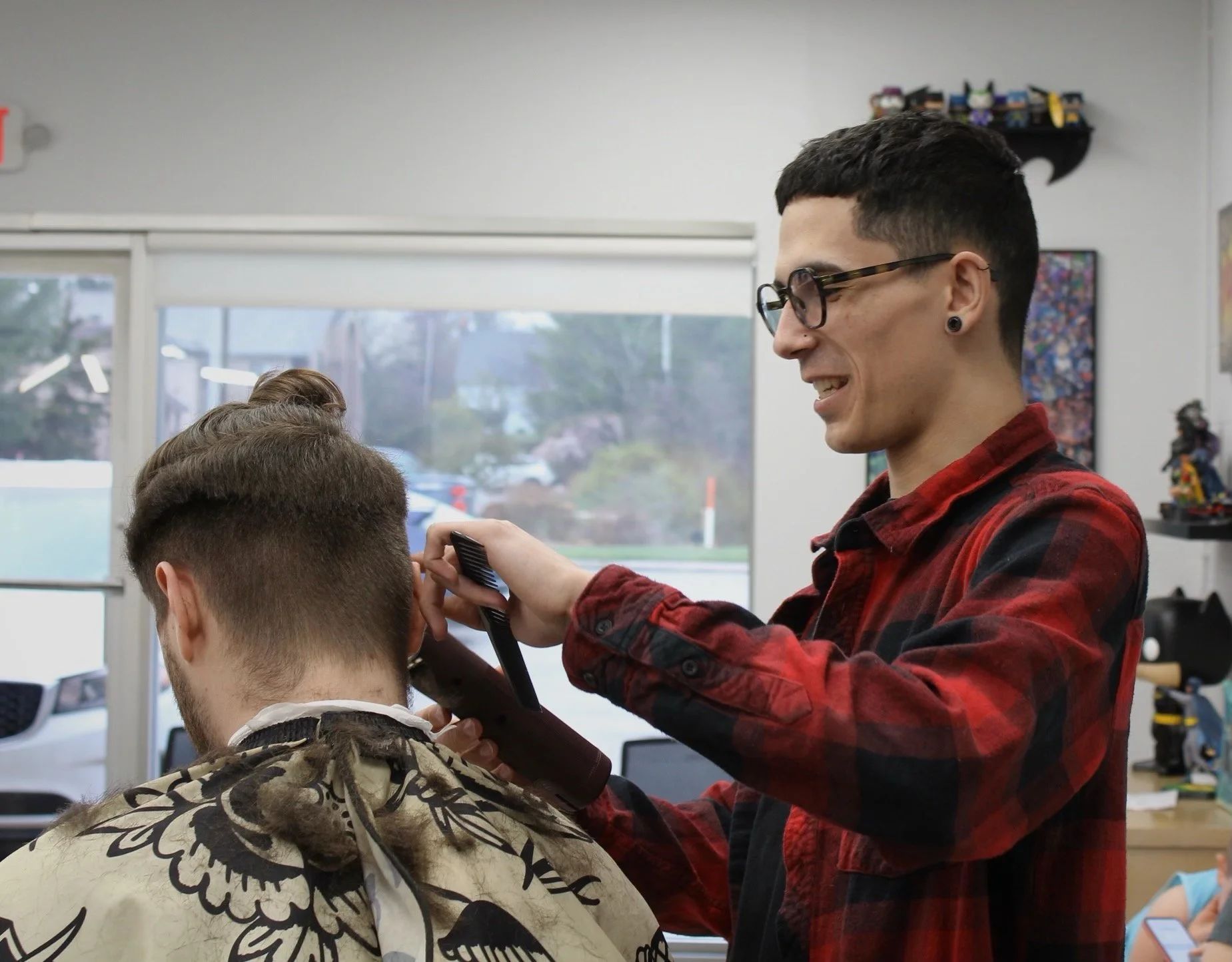 Barber giving a haircut in a shop. Man in red plaid shirt, holding clippers.