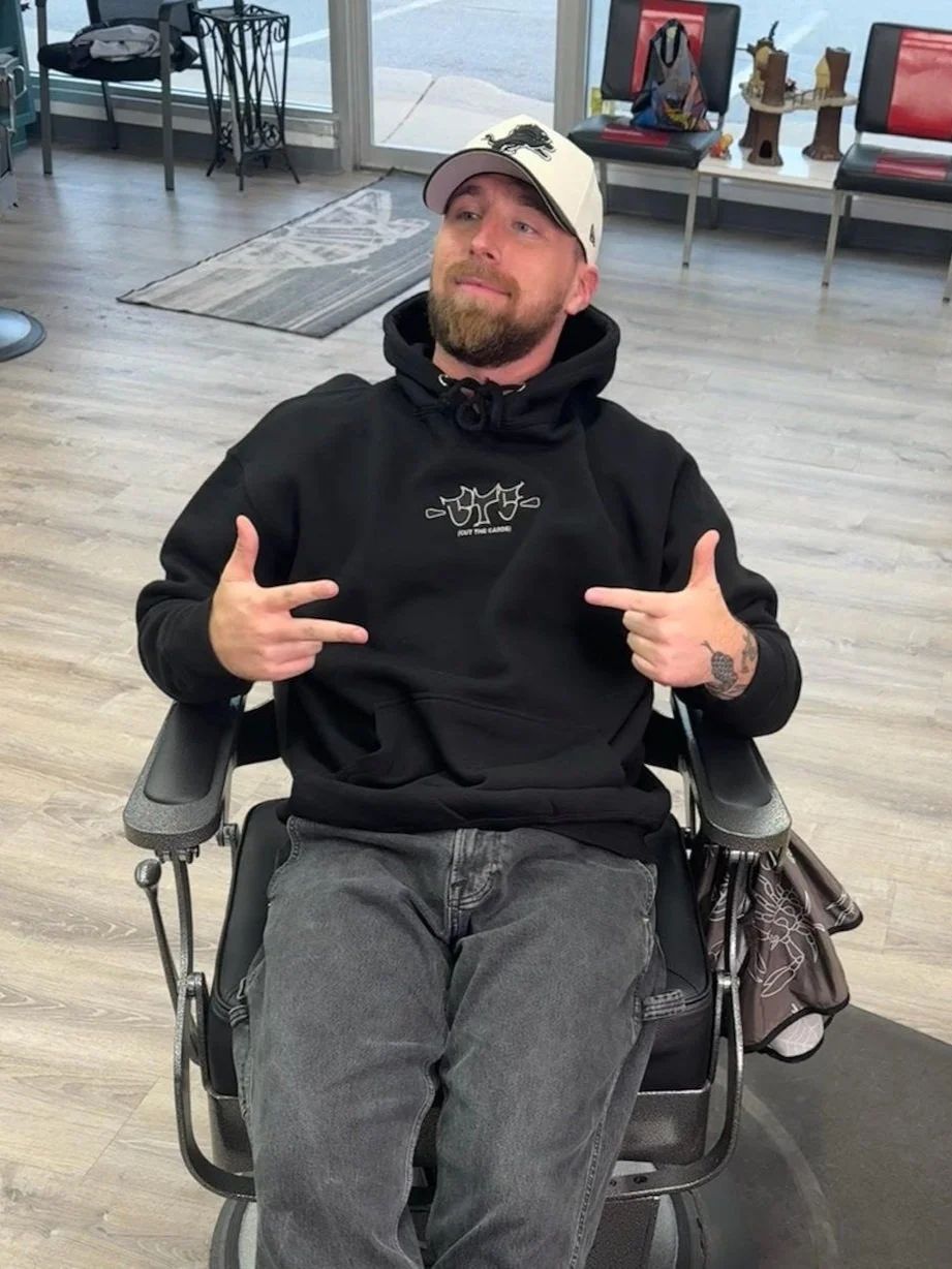 Man in a black hoodie and hat, sitting in a barber chair, pointing.