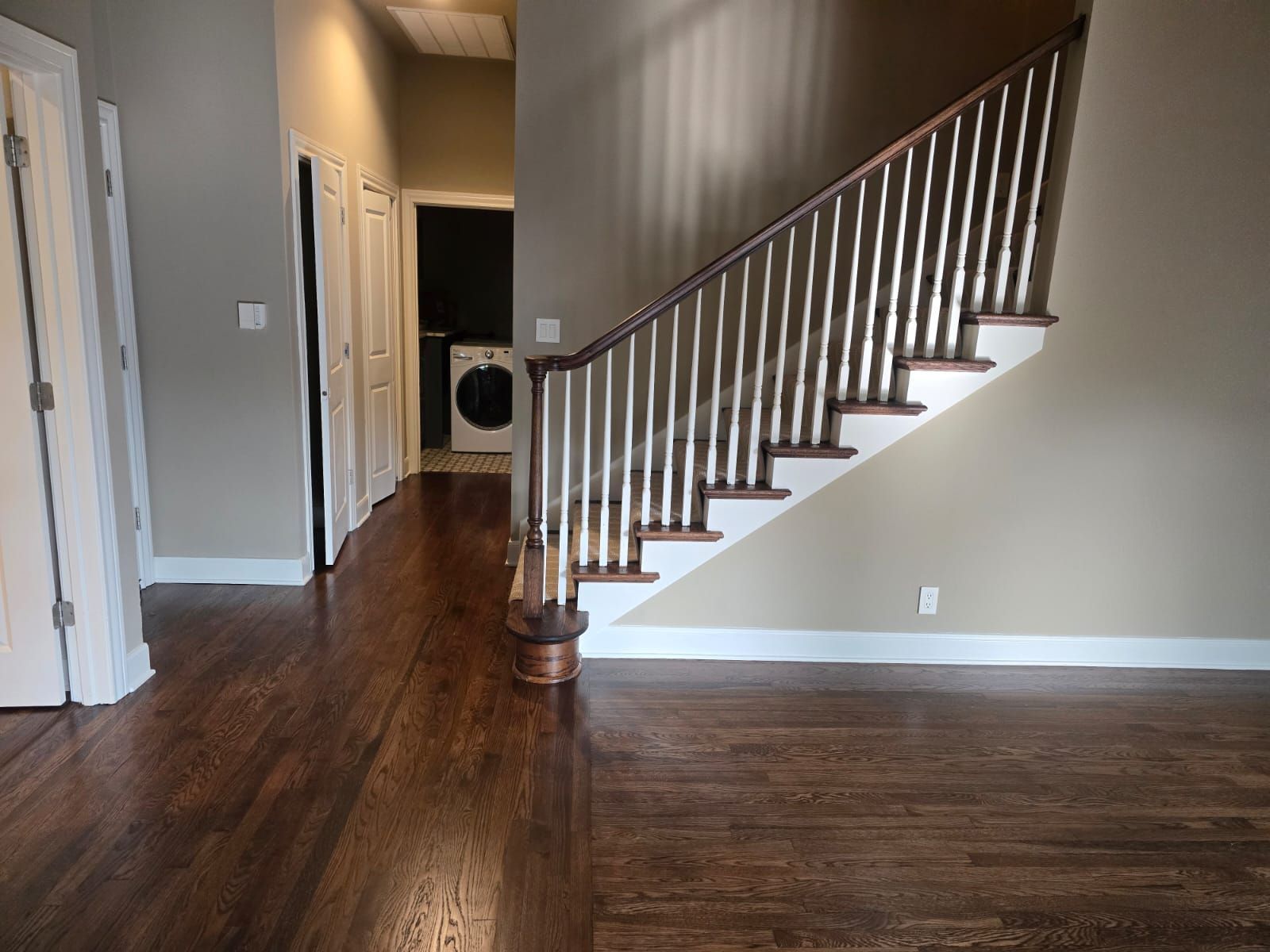 Dark wood floor with stairs and a hallway with a laundry machine visible. Brown wall with white trim.