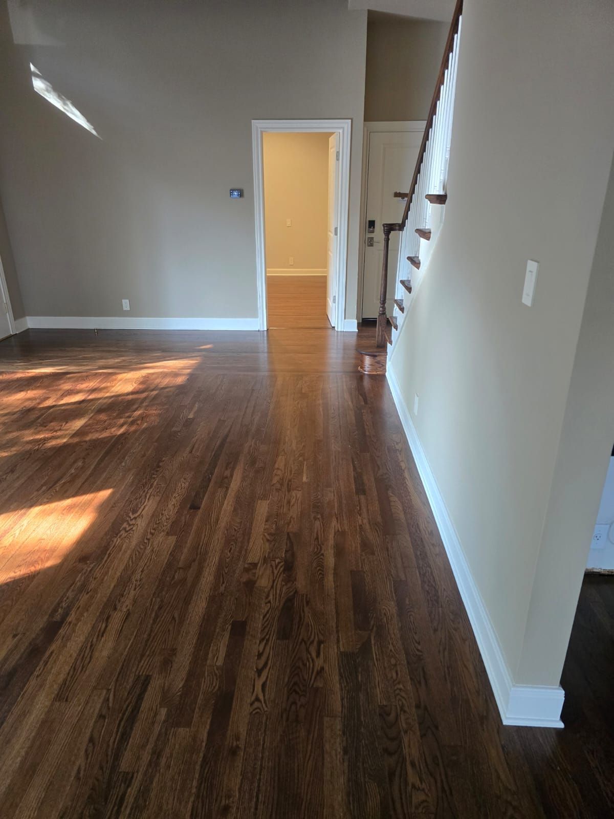 Hardwood floor in a room with a staircase, doorway, and tan walls. Sunlight illuminates the space.
