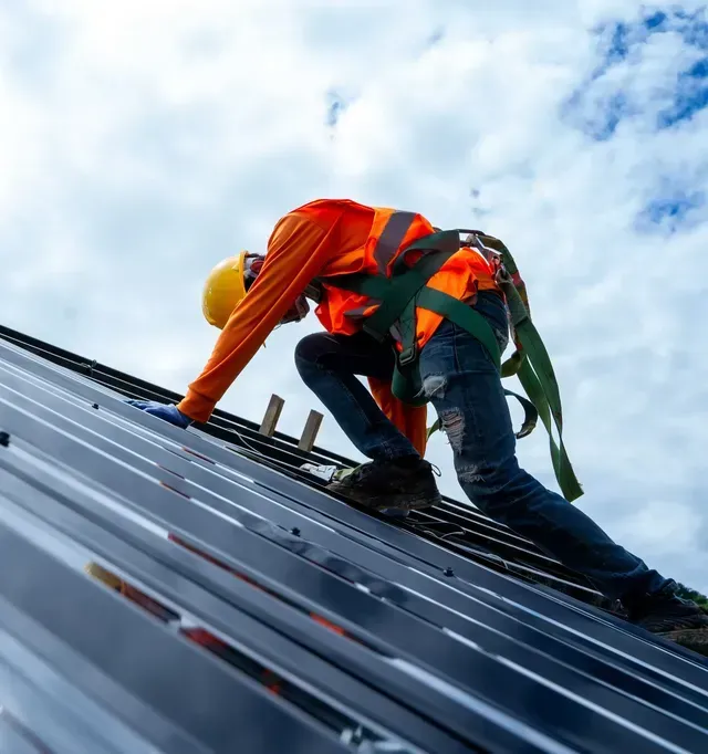 Roofer in hard hat and vest, hammering metal gutter on a rooftop. Sunset.