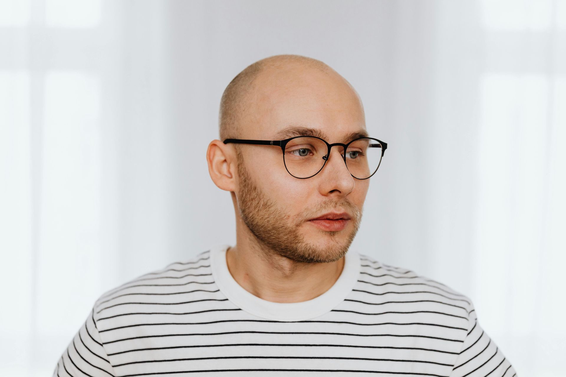 Bald person wearing glasses and a striped shirt, looking to the side in front of a white curtain.