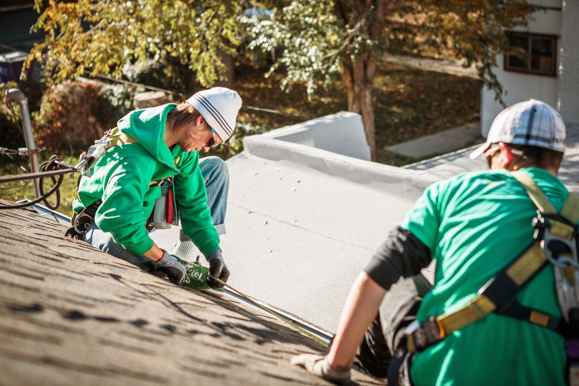 Construction worker on rooftop using a drill. He wears a white vest, hard hat, and holds a power tool.