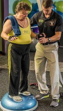 A man is helping an older woman stand on a balance ball in a gym.