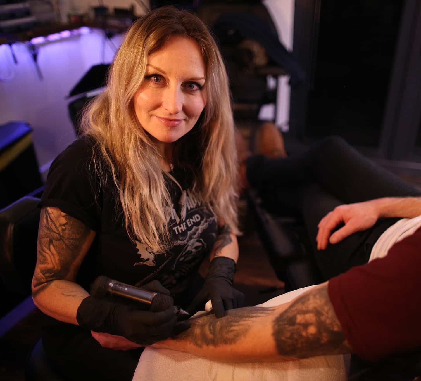 Helen tattooing a person's arm. She wears a black shirt and gloves, smiling as she works in a studio.