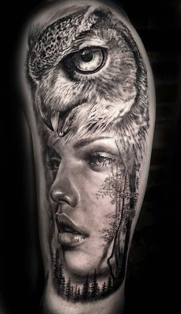Tattoo of an owl's head atop a woman's face, with forest trees visible on the side, grayscale.