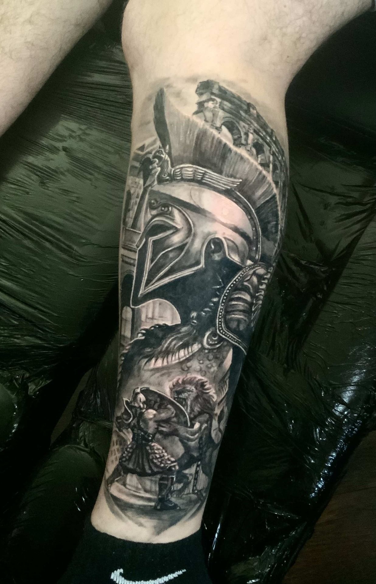Black and gray leg tattoo featuring a Spartan helmet and figures on a leg.