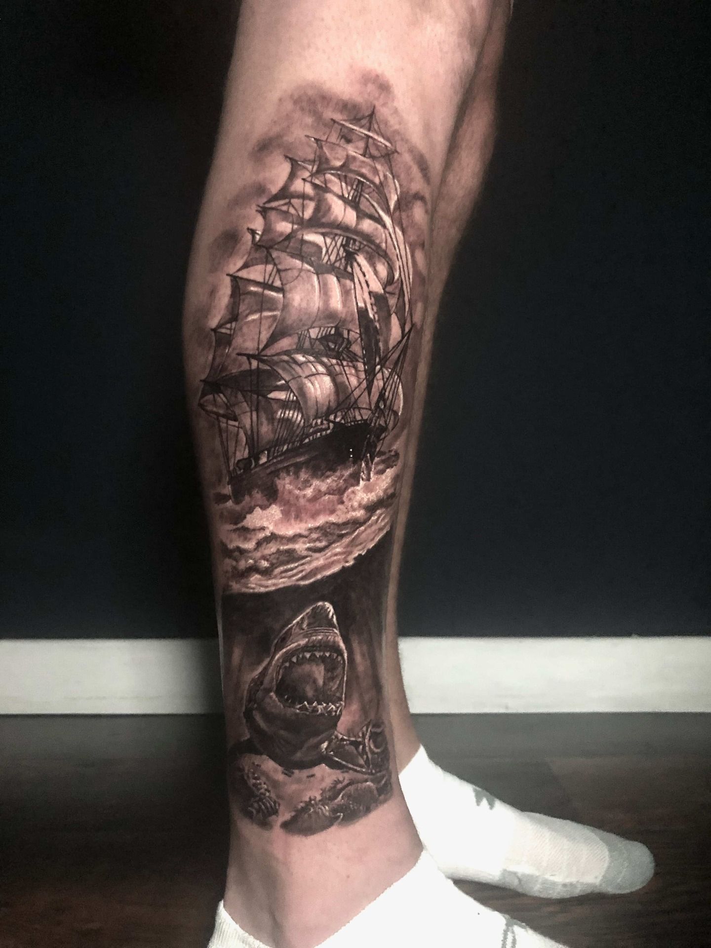 Tattoo of a sailing ship on turbulent waves, above an open-mouthed shark on a leg.