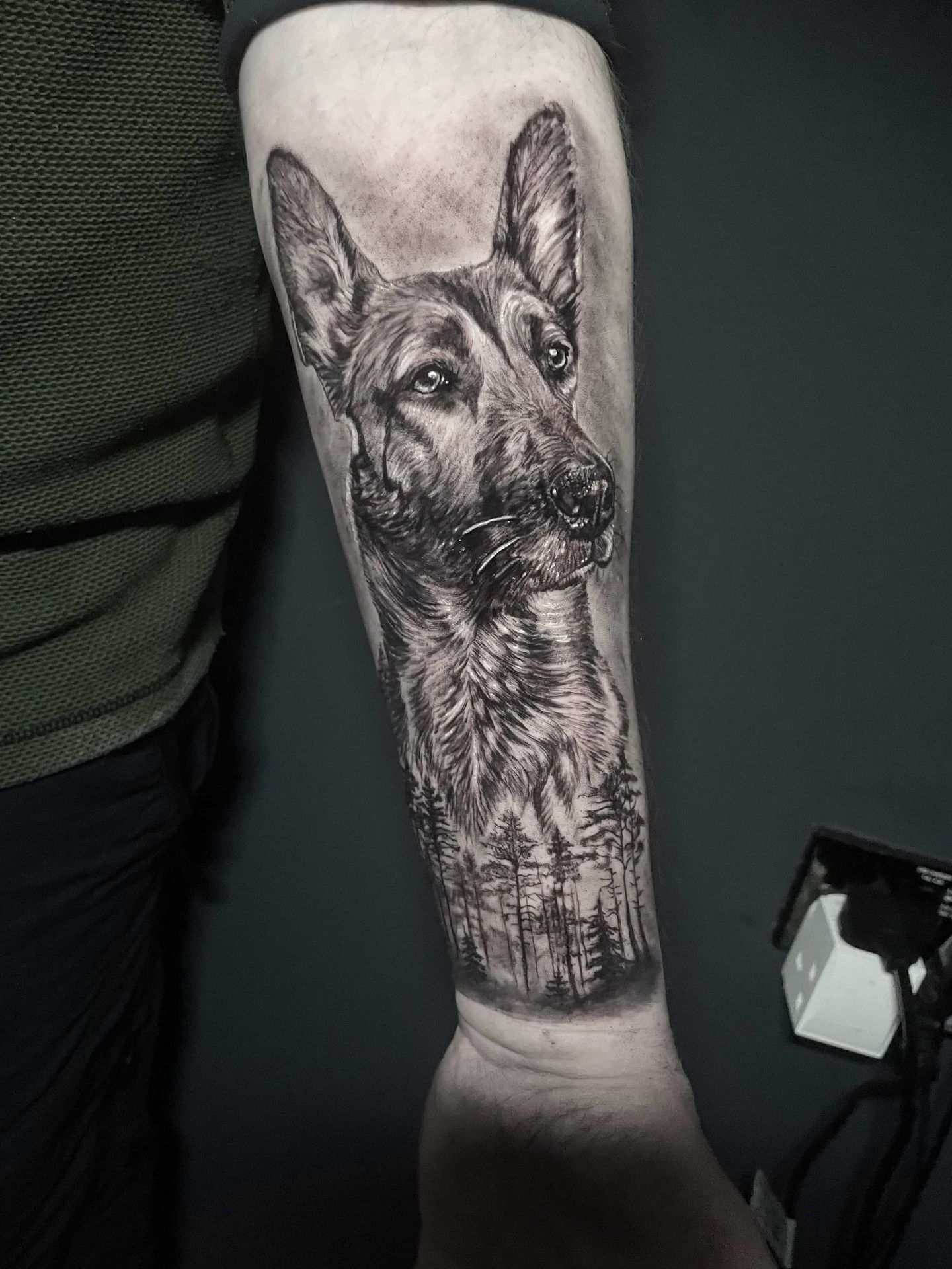 Black and gray tattoo of a German Shepherd on a forearm, with a forest scene at the bottom.
