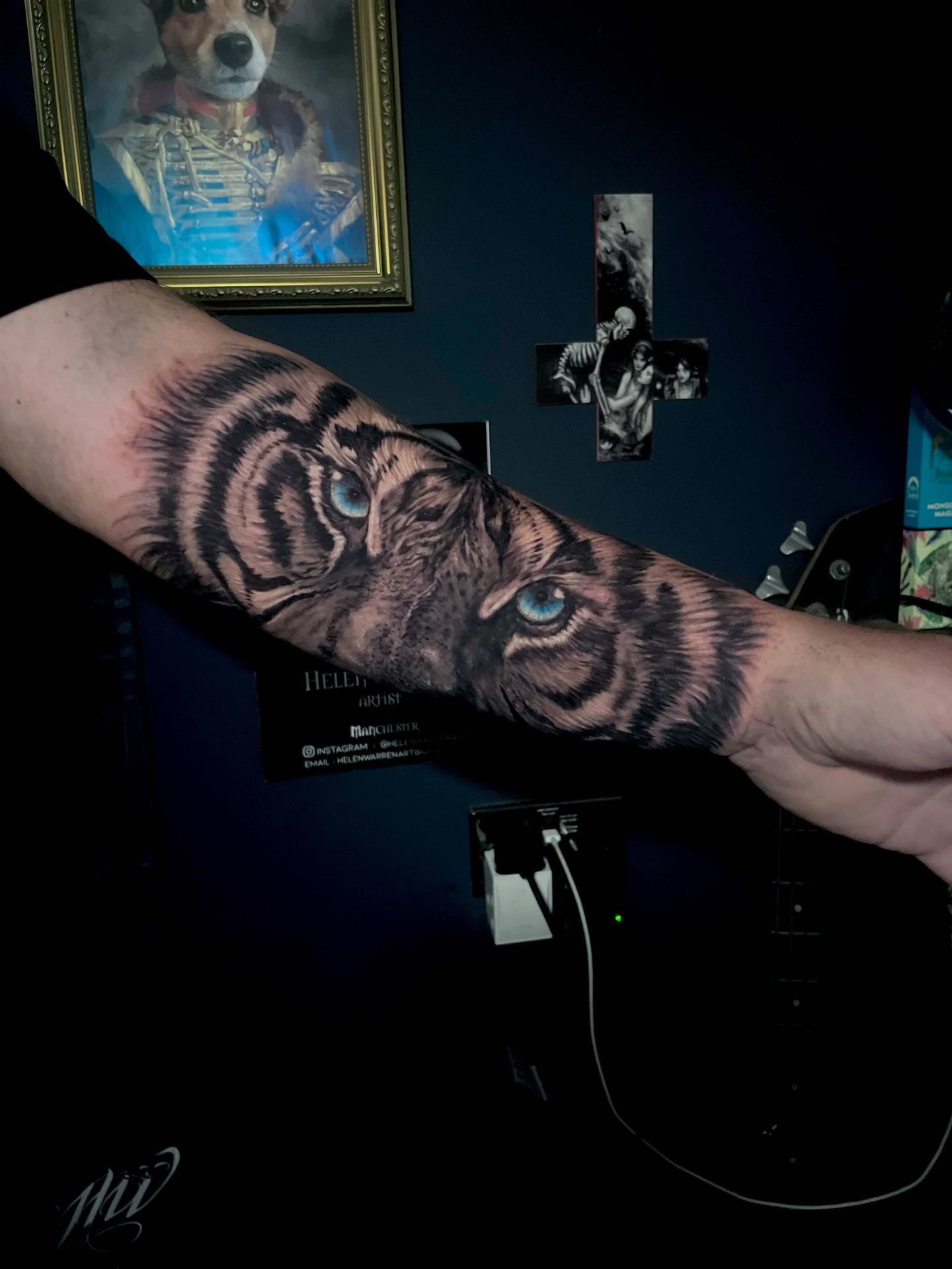 Tattoo of a tiger's face with blue eyes on a person's forearm; dark background.