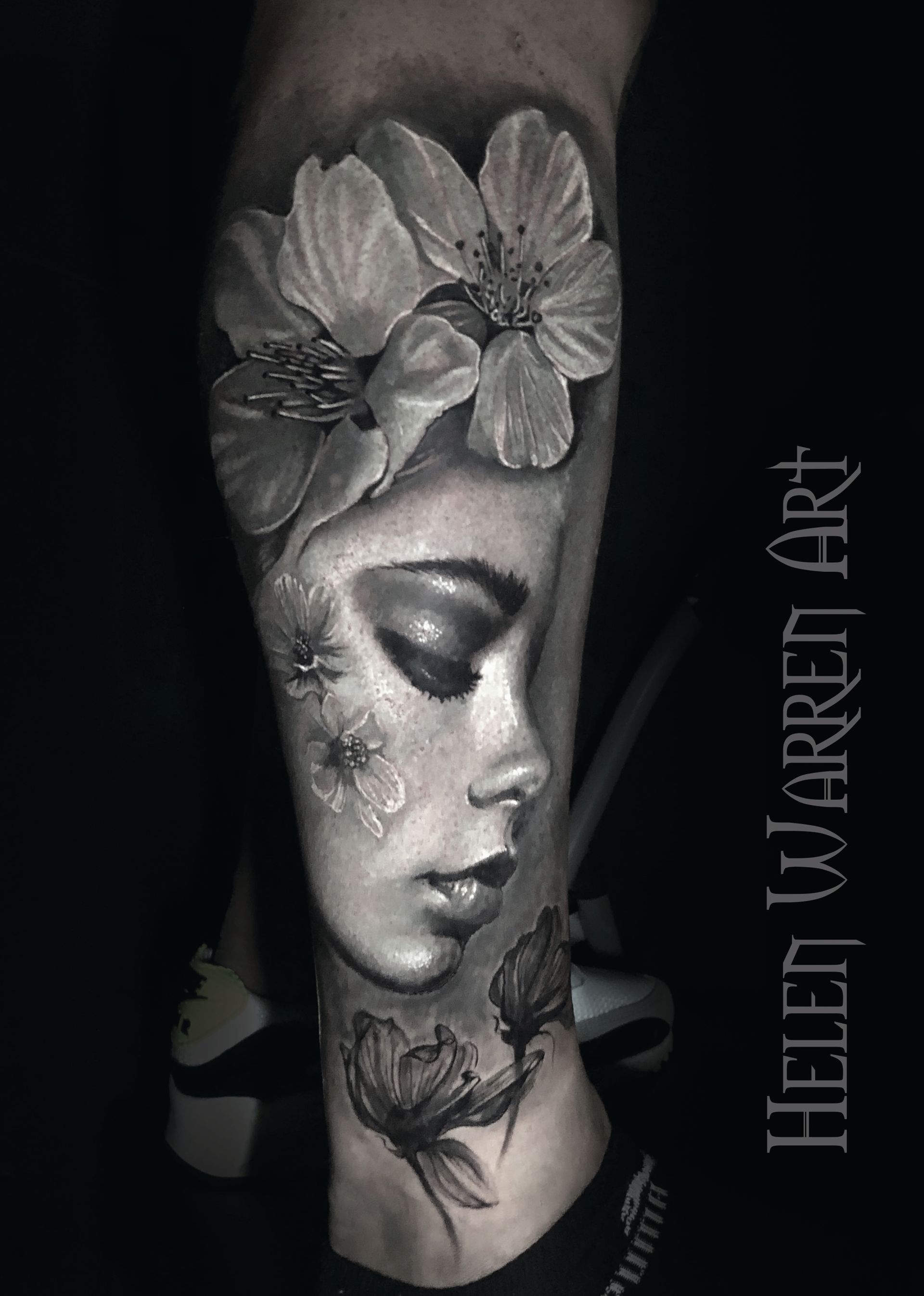 Black and white tattoo of a woman's face adorned with flowers on a leg, Helen Warren Art.