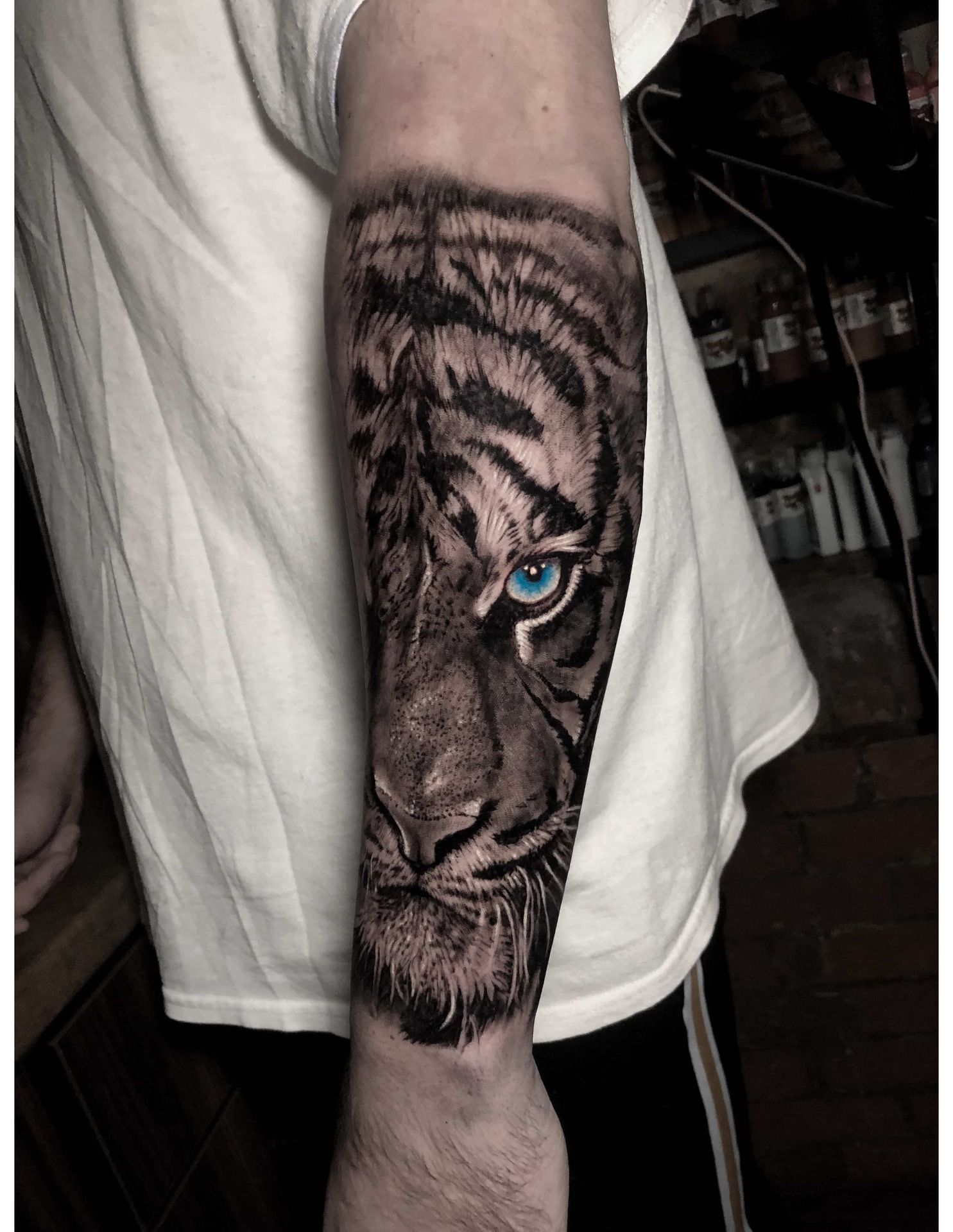Tattoo of a tiger's face on a forearm. Black and grey shading with a blue eye.