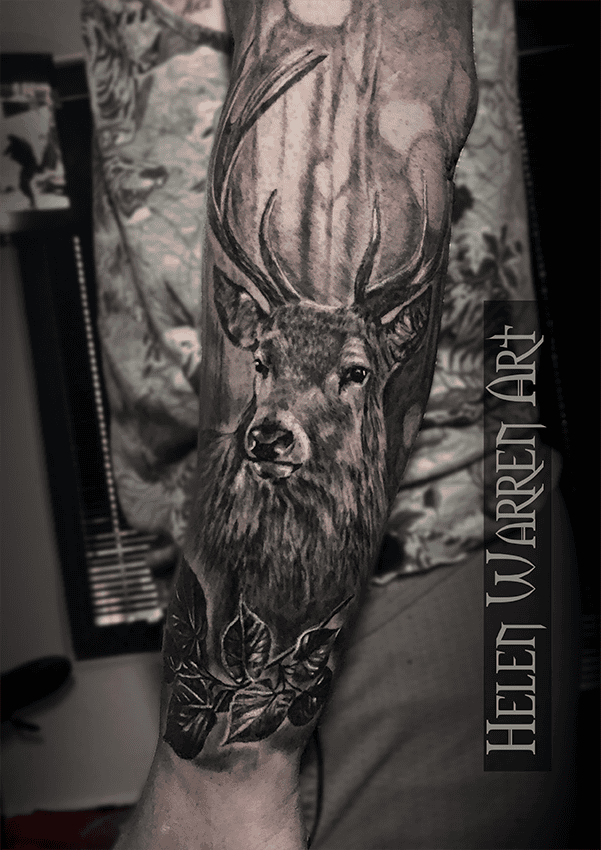 Black and white tattoo of a deer with antlers on an arm, forest background, leaves below.
