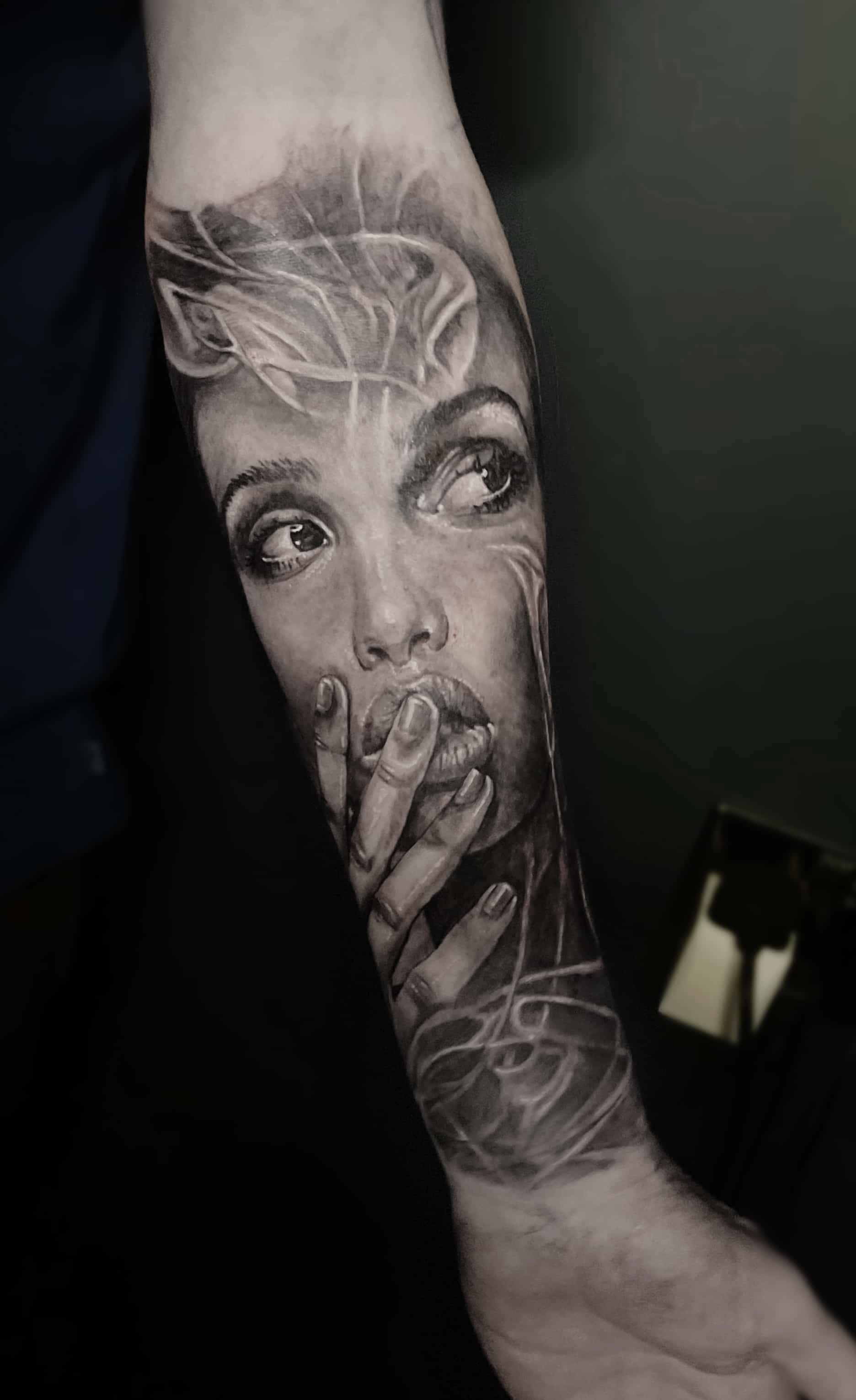 Black and white tattoo of a person’s face, holding hand near mouth, surrounded by wisps, on forearm.