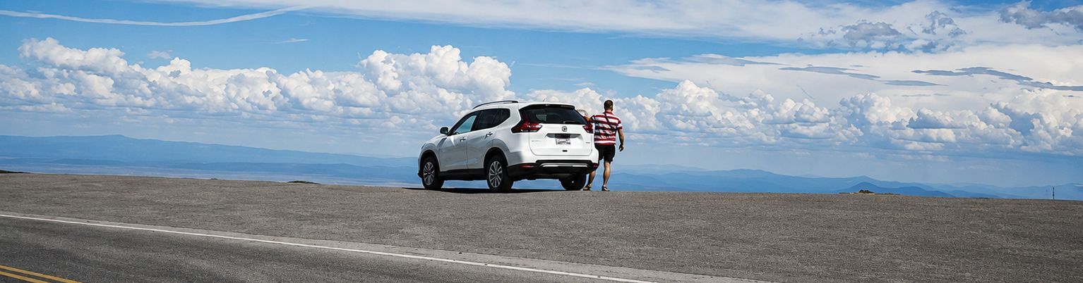 White SUV with a person standing beside it on a rocky shore, mountains and clouds in the distance