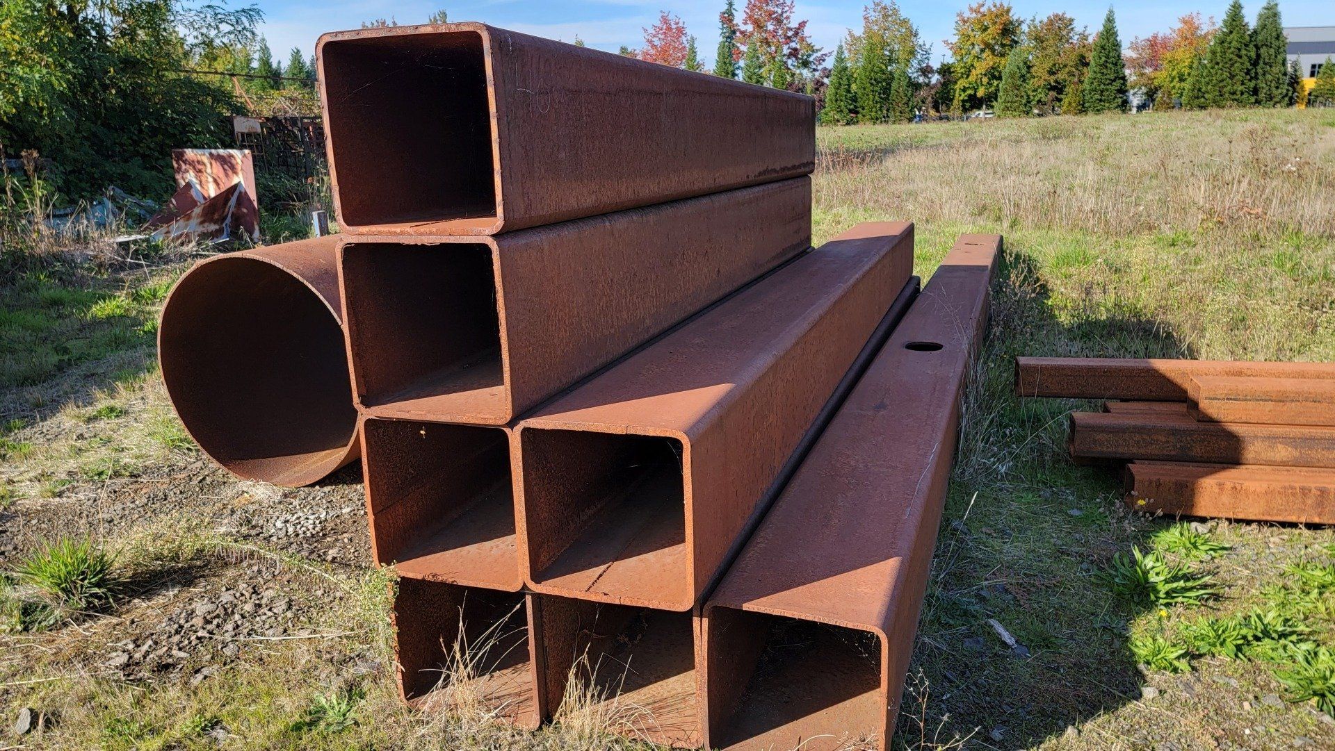 Rusty metal beams and a cylindrical pipe sit on a grassy field with a blurred background of trees and buildings.