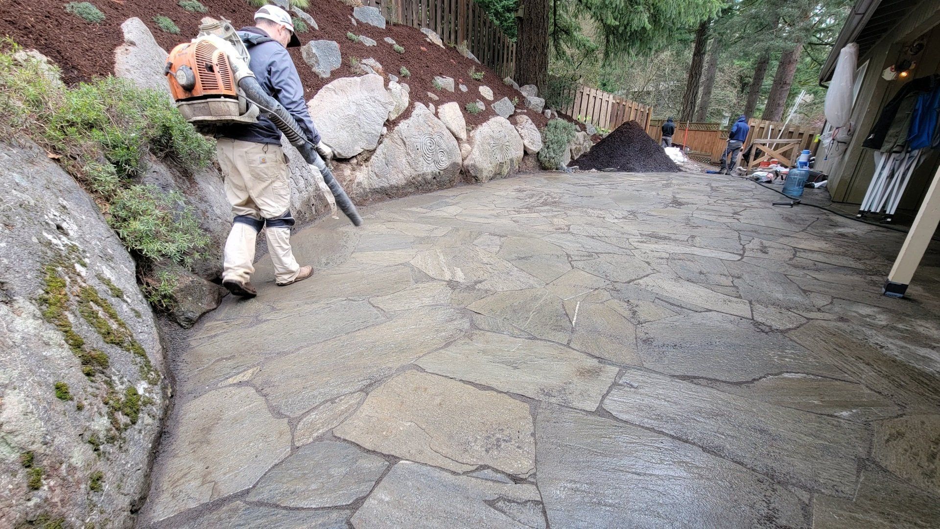 A person uses a leaf blower on a stone path, near a rocky hillside and a building.