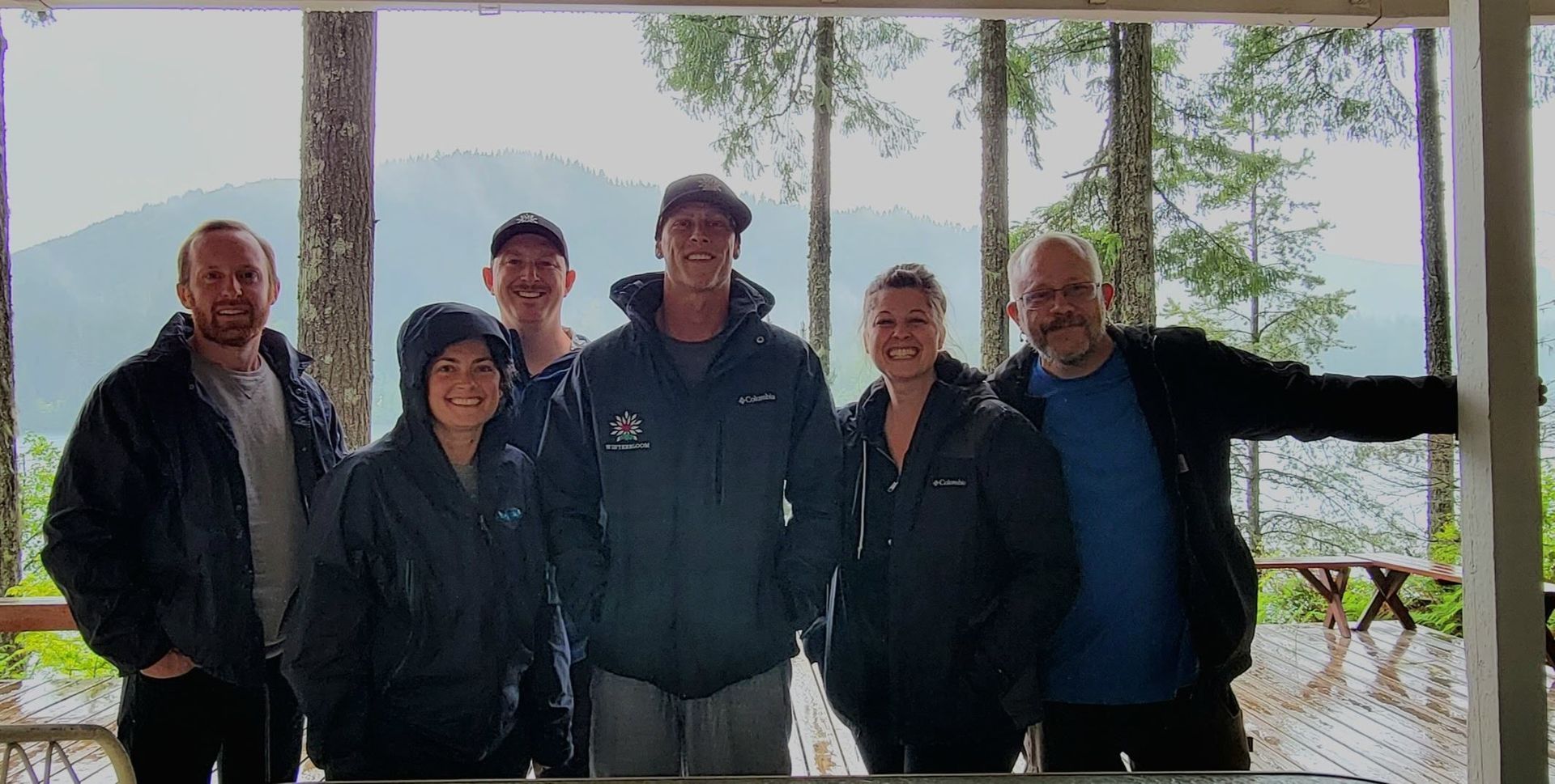 Group of seven people standing on a wooden deck with a scenic mountain and lake backdrop.