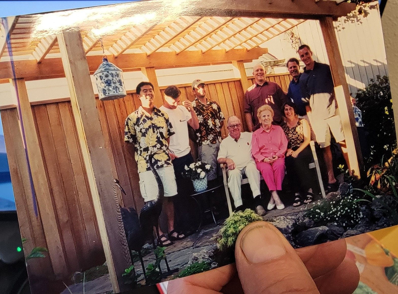 Family group posing outdoors under a wooden pergola, including older couple seated in front and others standing; sunny day.