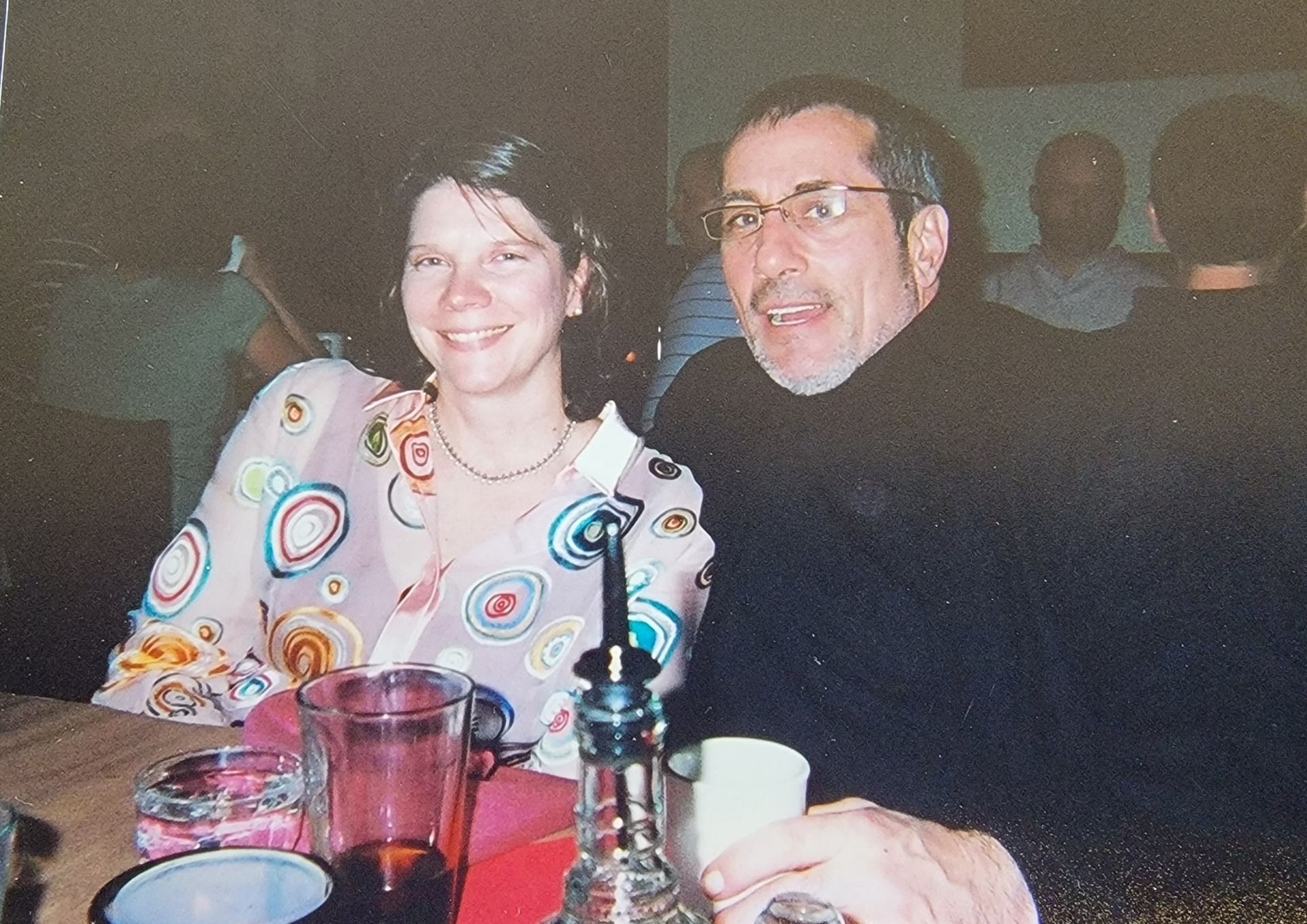 Woman and man smiling at a table. The woman wears a colorful patterned blouse. 