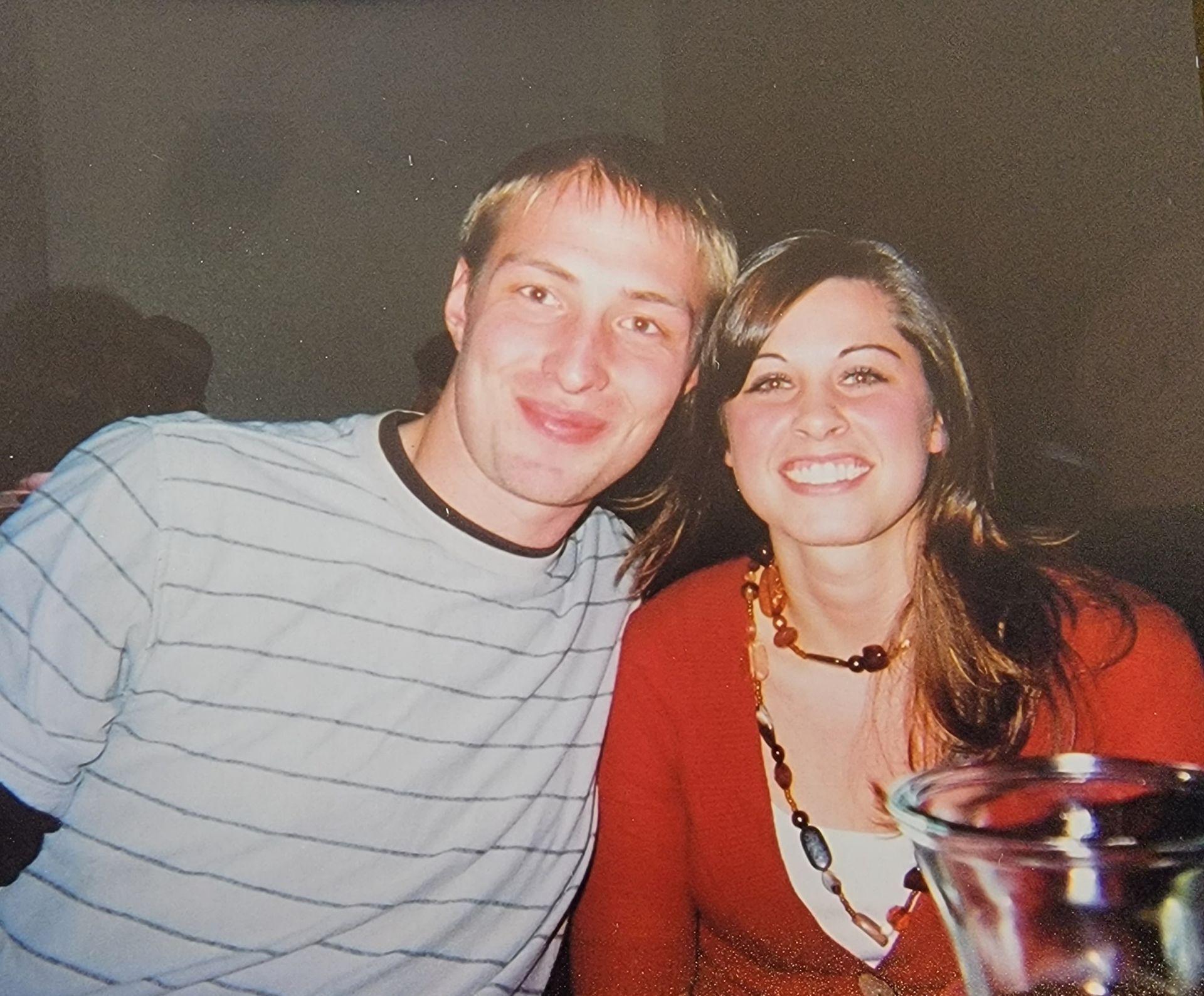 Smiling couple posing close together indoors. The woman wears a red shirt and necklace. The man has a striped shirt.