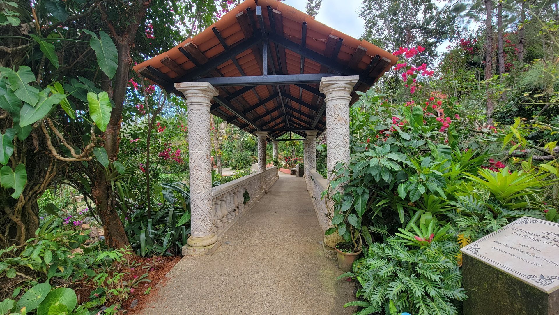 Covered walkway with stone columns and red-tiled roof, surrounded by lush greenery.