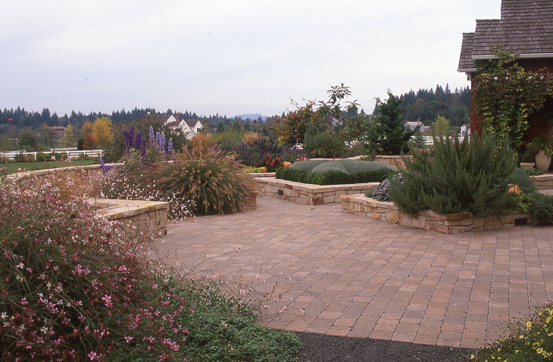 Brick patio with raised stone planters filled with colorful flowers and greenery; a house is in the background.