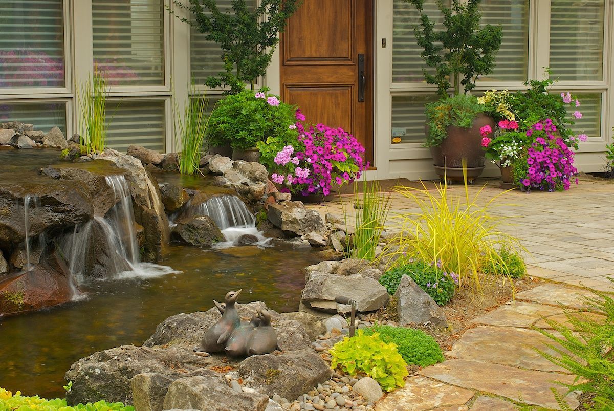 A small water feature with a waterfall and pond in front of a house with flowers and a walkway.