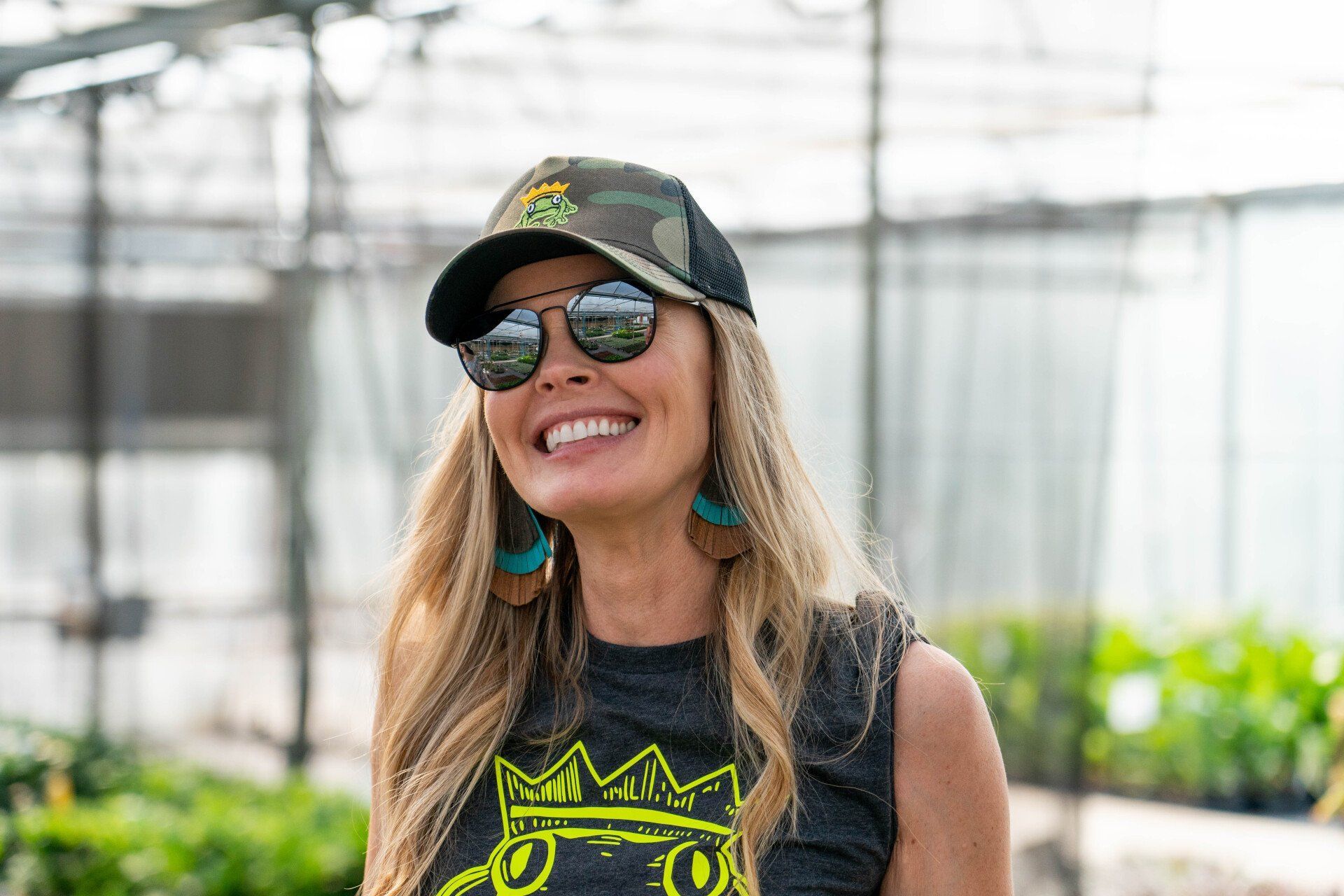 Woman with blonde hair and sunglasses smiles in a greenhouse, wearing a camouflage hat and earrings.