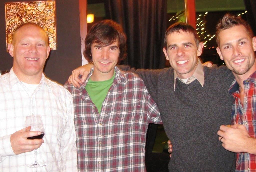 Four men smiling, posing indoors. One man holds a wine glass. They wear casual clothing.