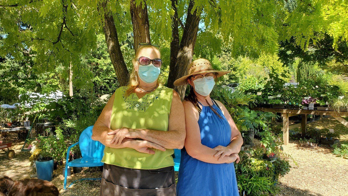 Two women wearing masks stand under a tree with arms crossed. 
