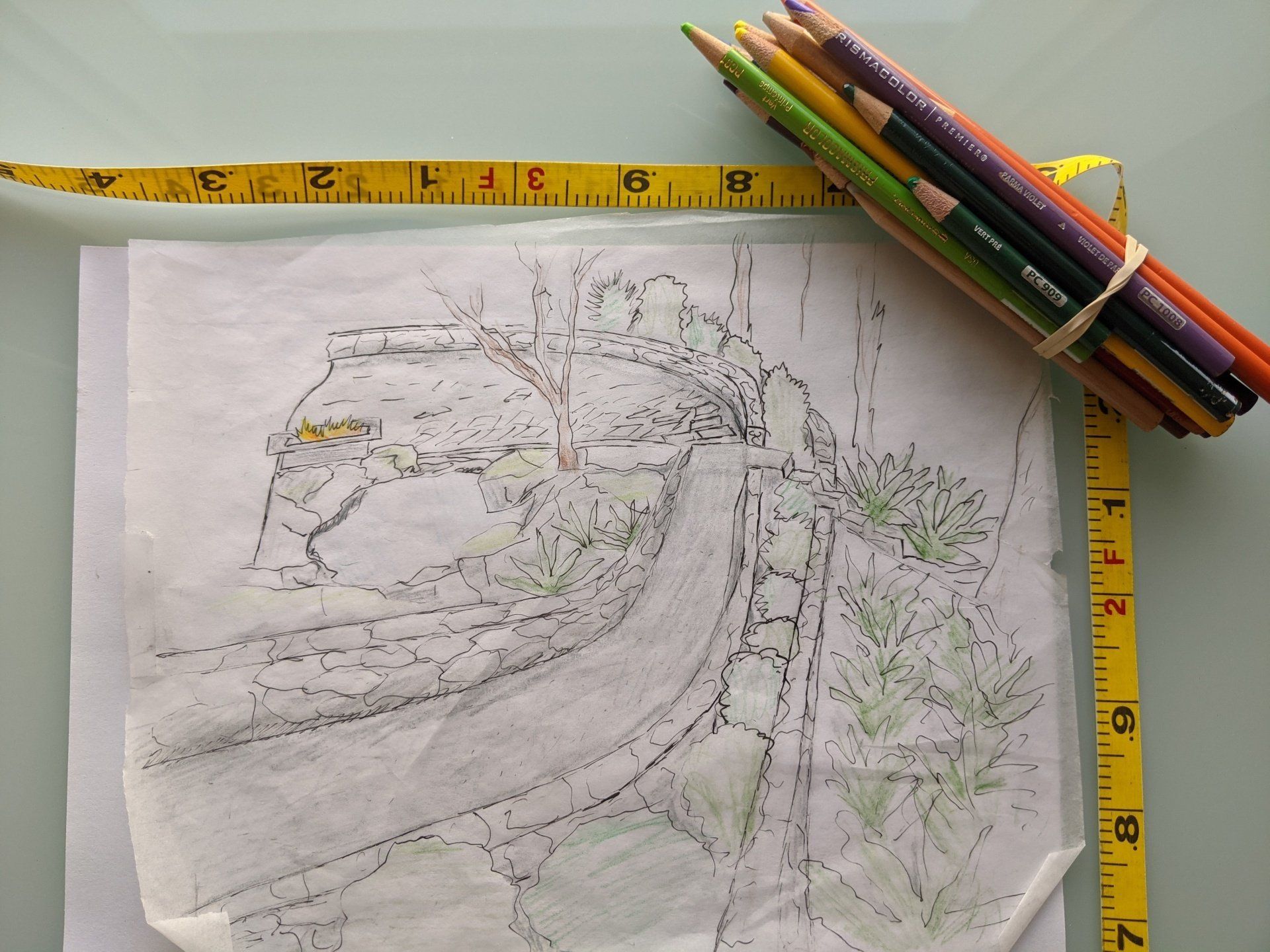 Sketch of a bridge and plants, partially colored with green, beside a yellow measuring tape and a bundle of colored pencils.