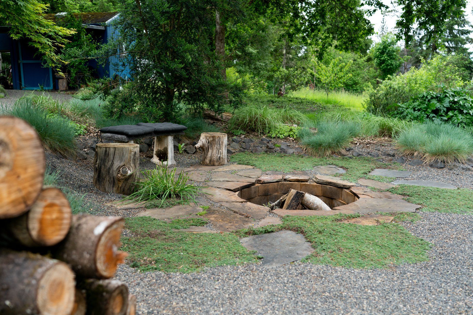 Fire pit area with log seating, firewood stack, and surrounding greenery. 
