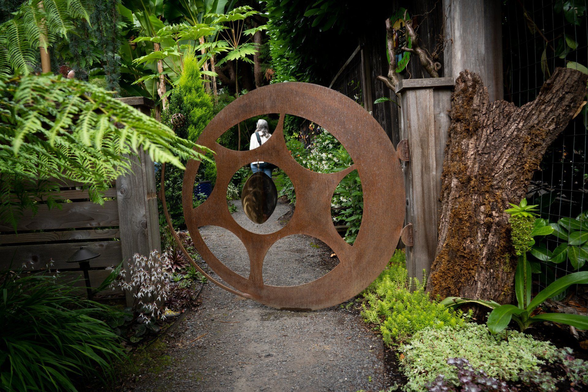 Rusty metal gear gate in a garden pathway entrance, surrounded by greenery and wooden posts.