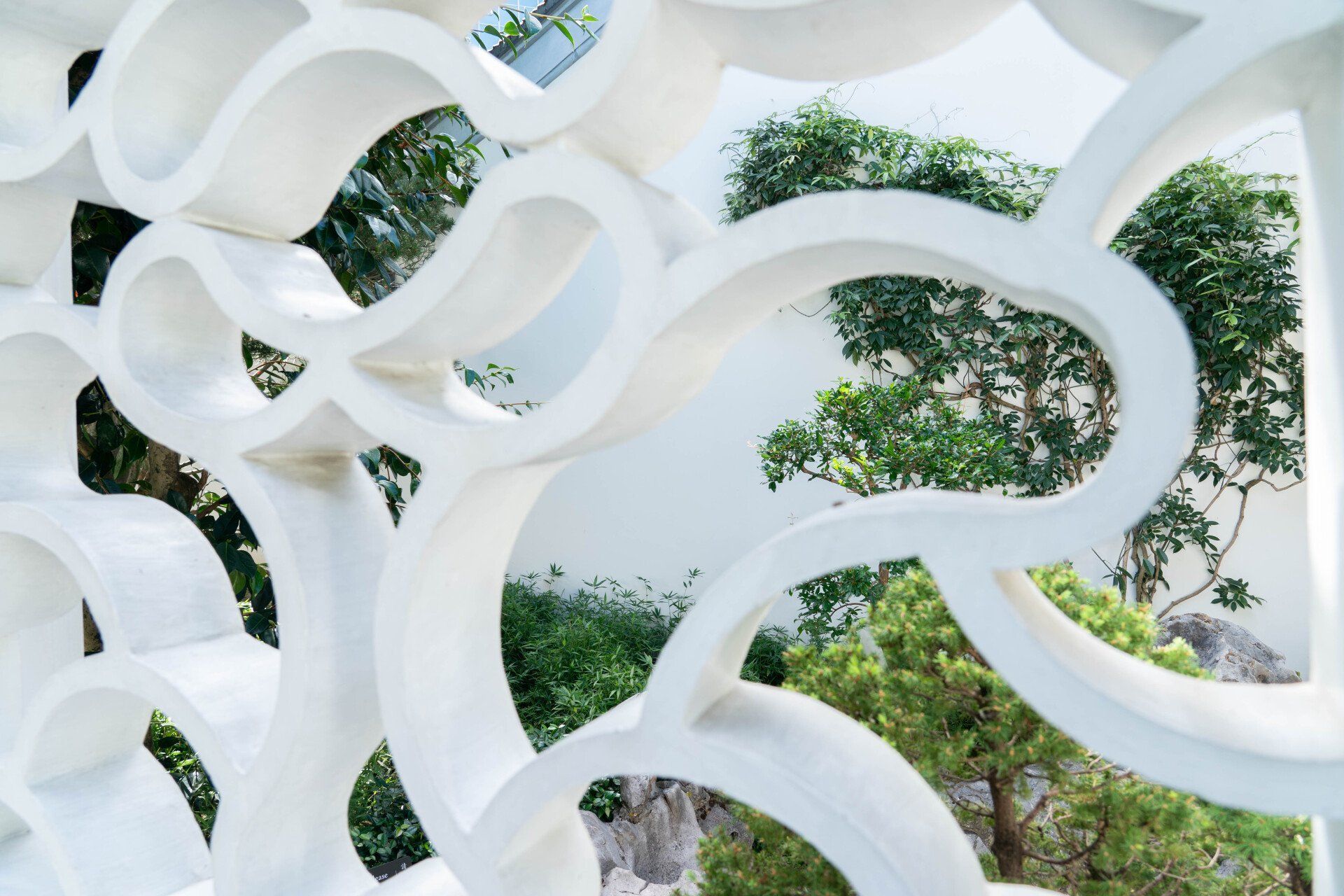 White, ornate, curved architectural screen framing a view of green trees and a white wall.