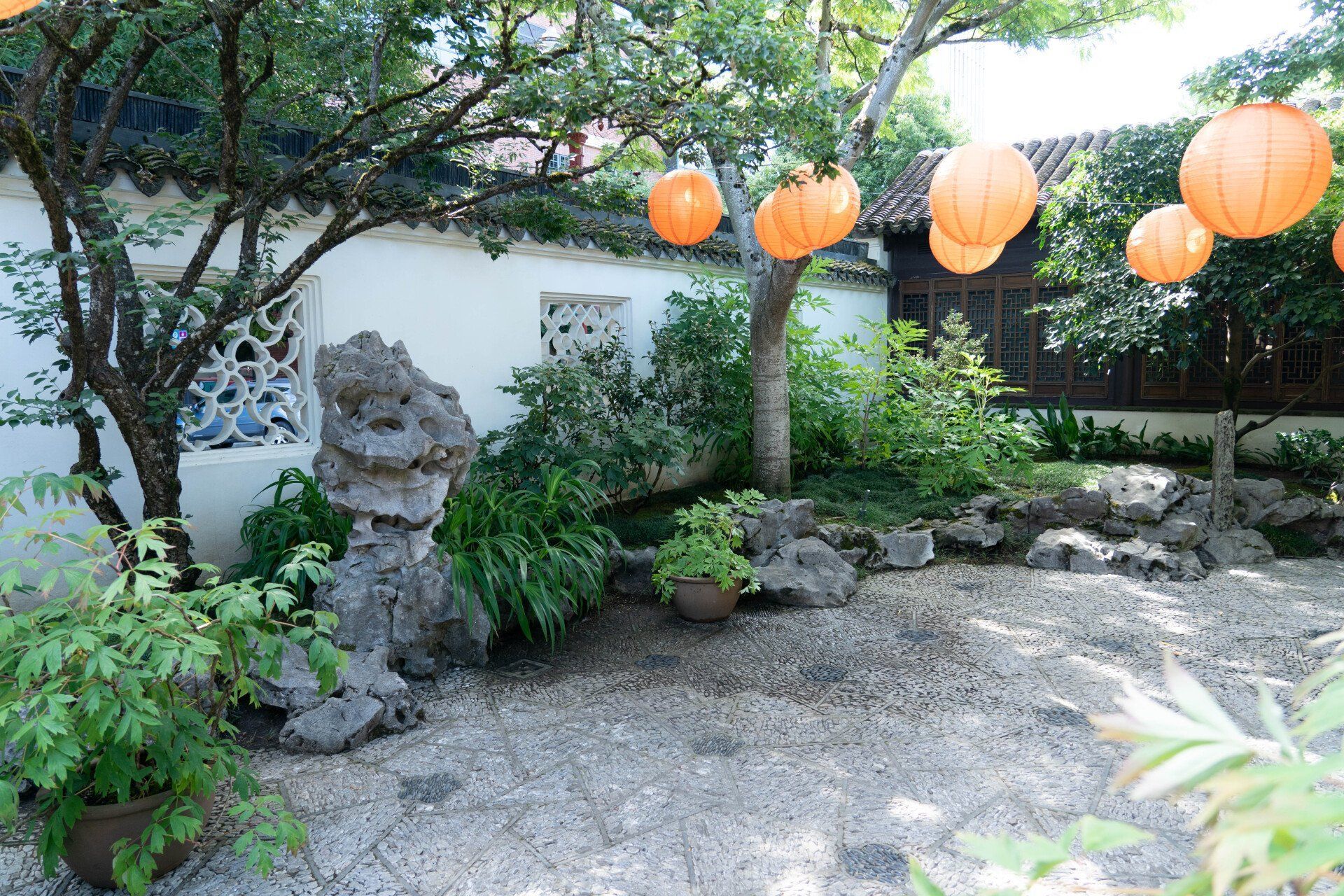 A serene Chinese garden with orange paper lanterns, stone features, lush greenery, and a white wall.