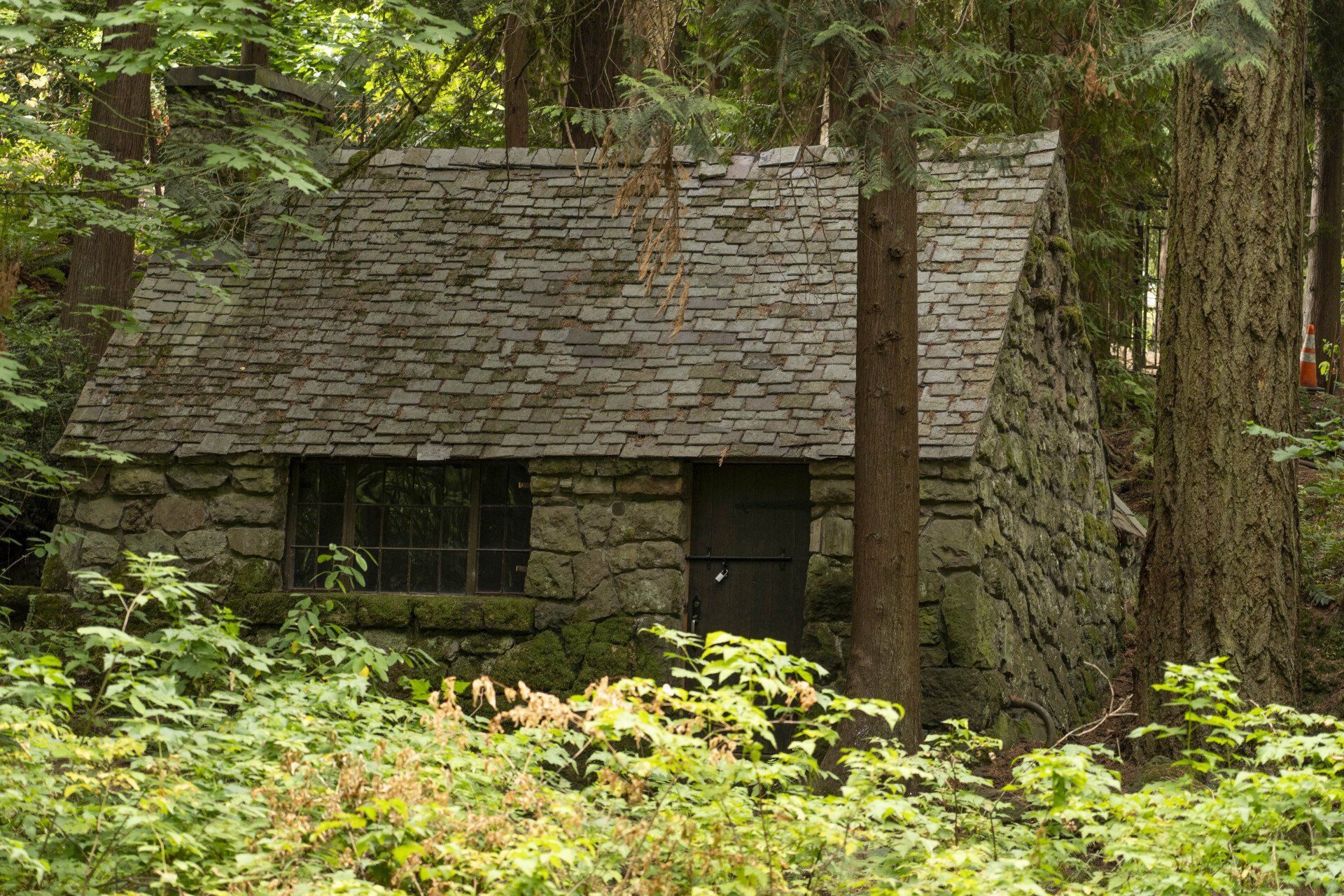 Stone cabin nestled in a lush forest, with a slate roof and small windows, surrounded by green foliage.