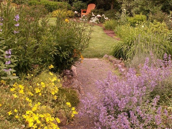 A garden path leads to a chair; yellow and purple flowers frame the path; green grass and shrubs surround it.