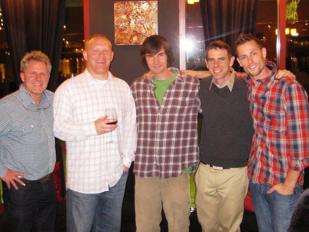 Five men pose indoors, arms around each other. 