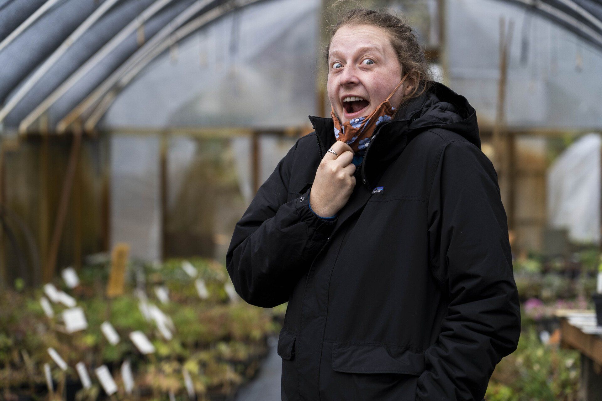 Person in a black jacket, mouth open, holds and bites a brown item in a greenhouse.