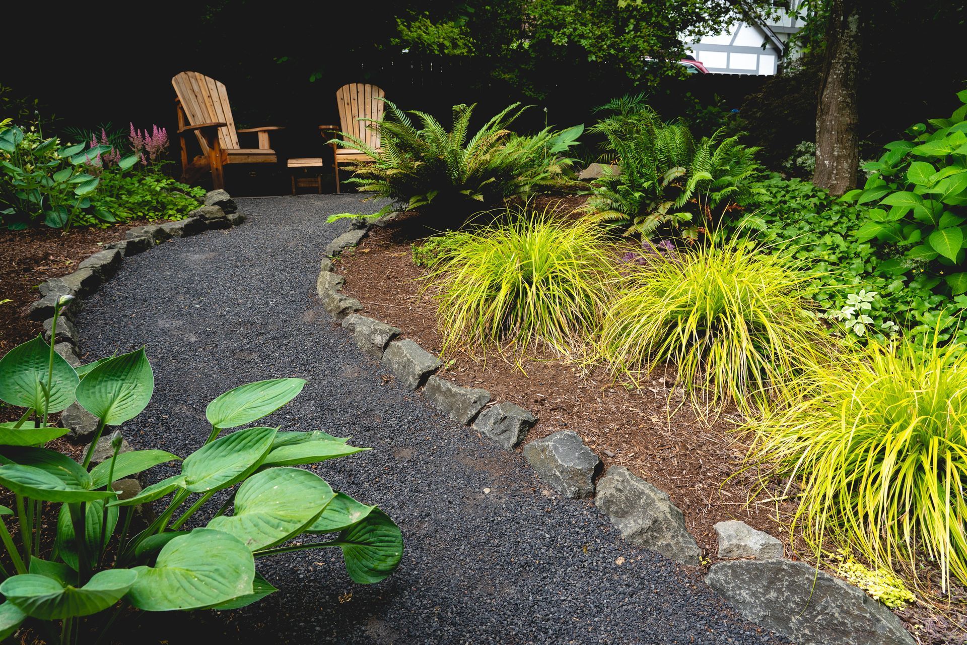 Gravel path in a garden with lush green plants, leading to two wooden chairs under a tree.