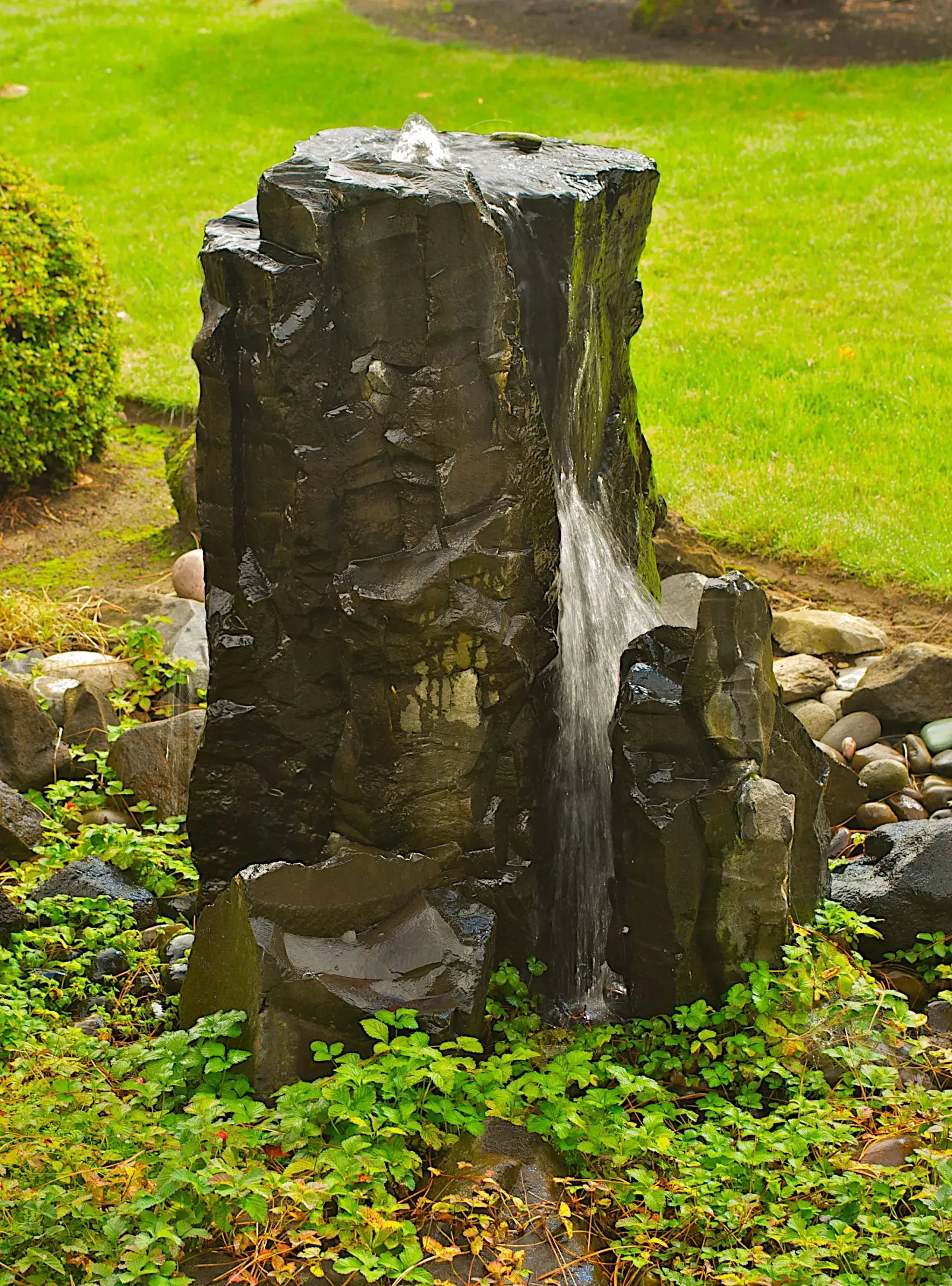 Stone water fountain in a garden setting, with water cascading down its dark, textured surface.