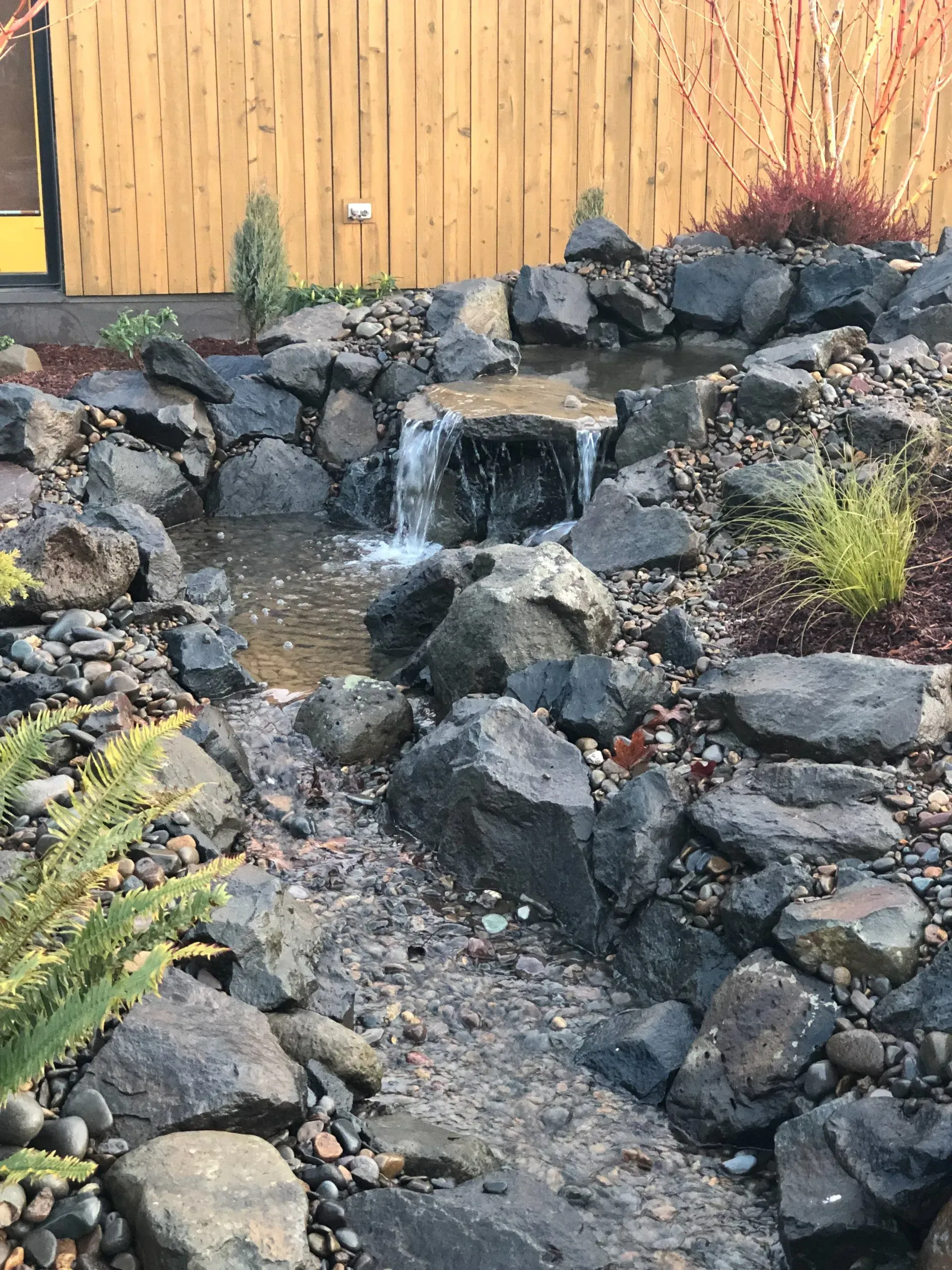 Small stone waterfall flowing into a pond, surrounded by dark rocks and greenery, near a wooden fence.