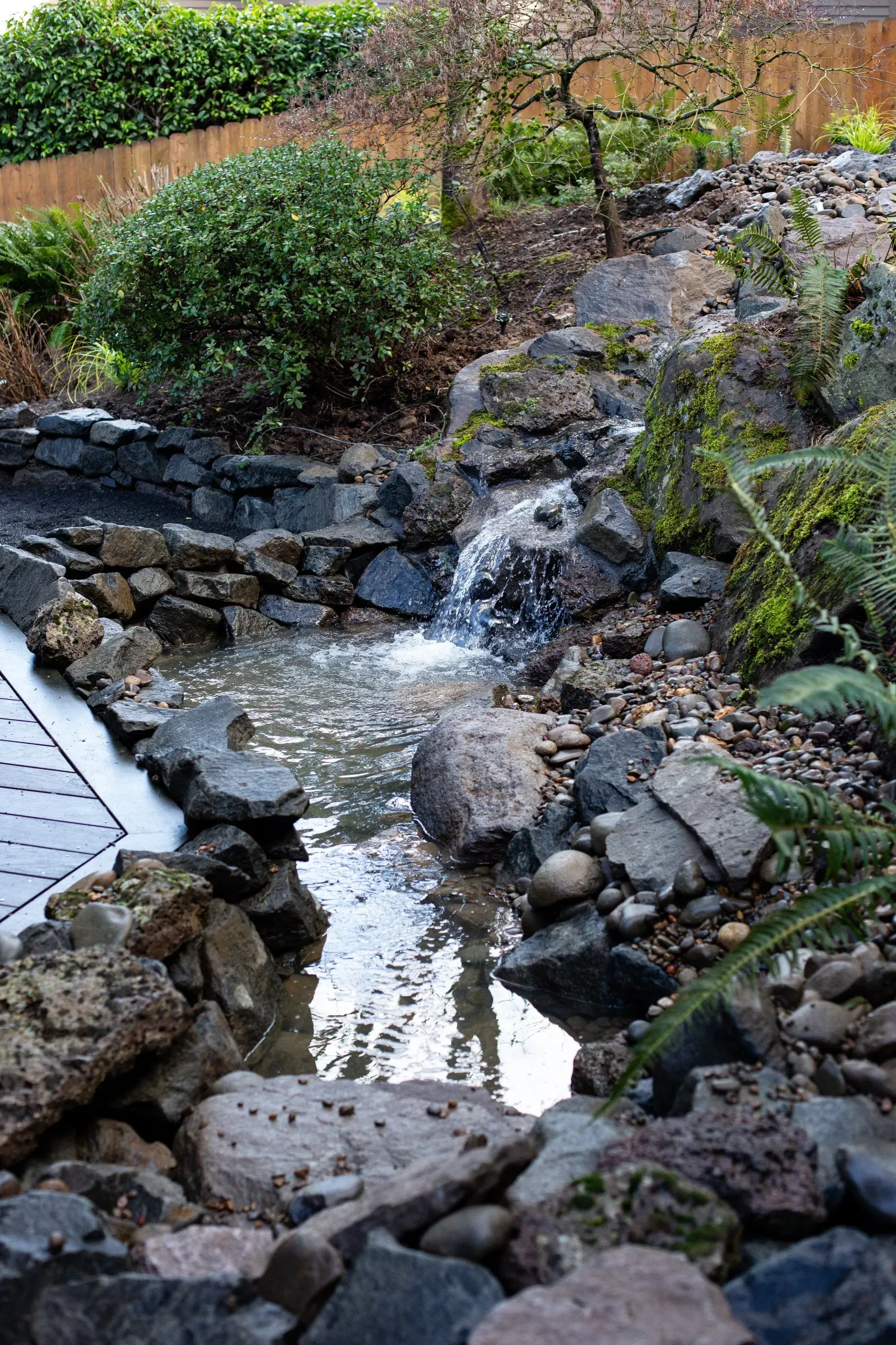 A rocky water feature with a small waterfall cascades into a pond, surrounded by greenery.