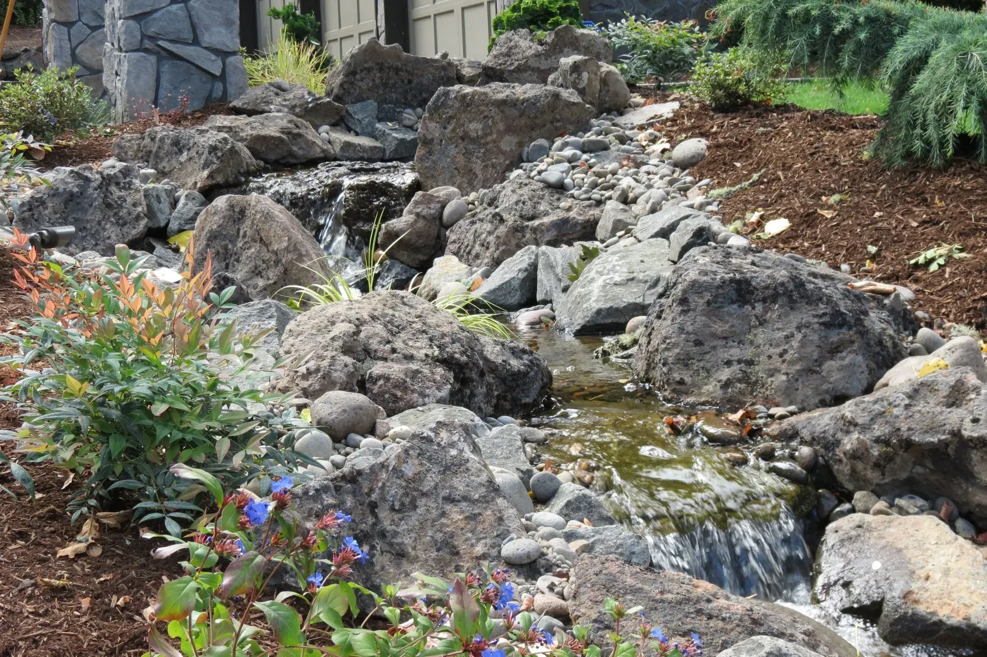 A small man-made stream flows through a rocky landscape with plants and a few small waterfalls.
