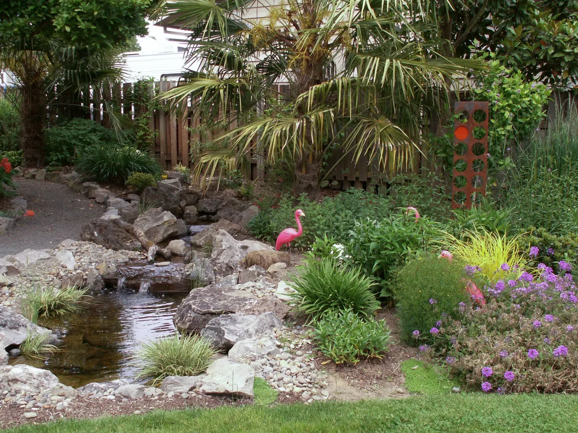 A garden scene with a small pond, rocks, lush greenery, and a pink flamingo statue.