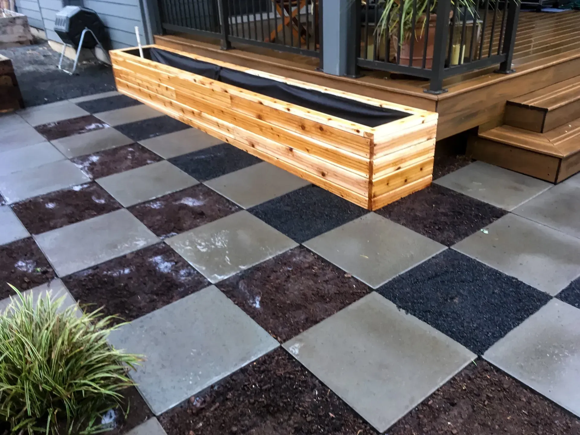 A checkerboard patio with gray, dark, and reddish-brown squares, next to a wooden planter box and deck.