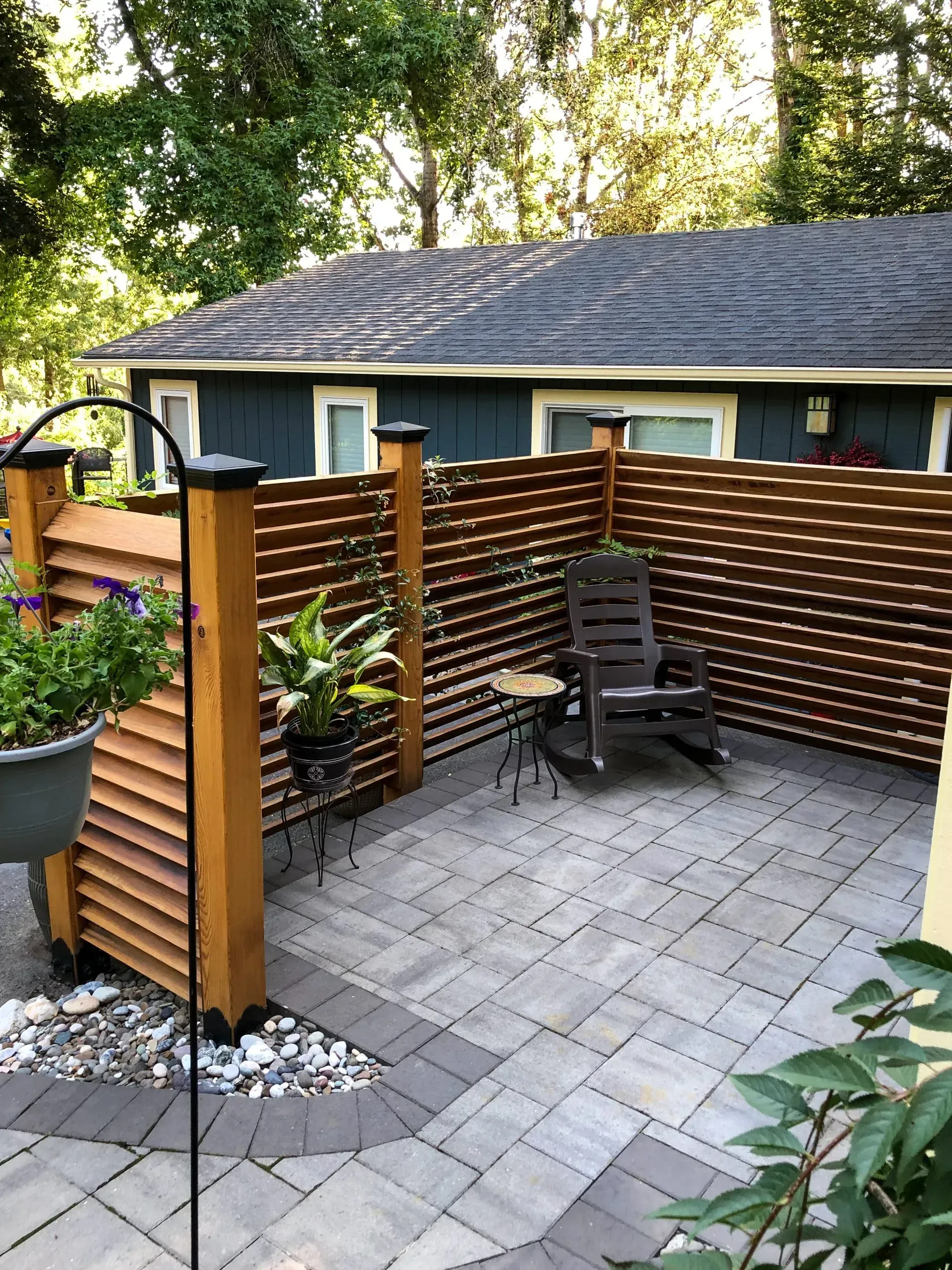 Cozy backyard patio with gray pavers, a wooden slatted privacy fence, and a rocking chair; a blue house is in the background.